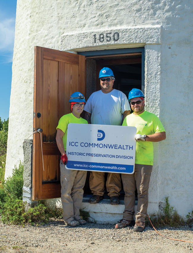 Three crew members in the light house door holding an CC Commonwealth sign