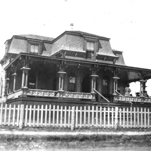 Black and white image of a house with wrap around porch and mansard roof